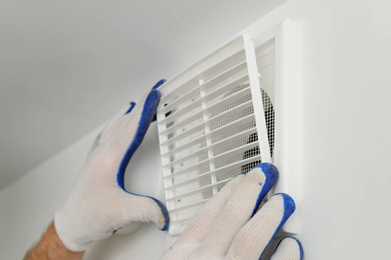 Main gantée tenant une grille d'aération blanche avec lamelles orientables et moustiquaire intégrée, fixation murale ou plafond visible