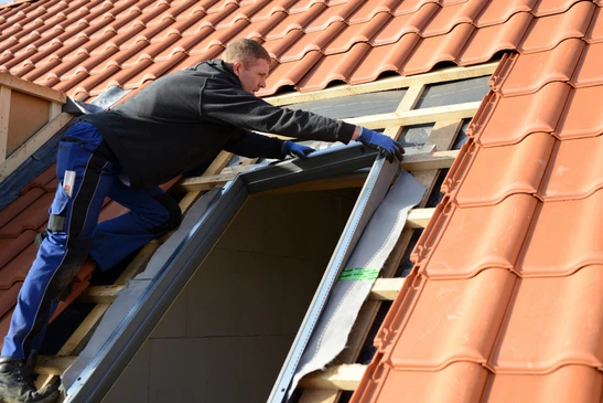 Un homme installe un film plastique de protection autour d'un châssis de fenêtre de toit Velux dans des combles en construction